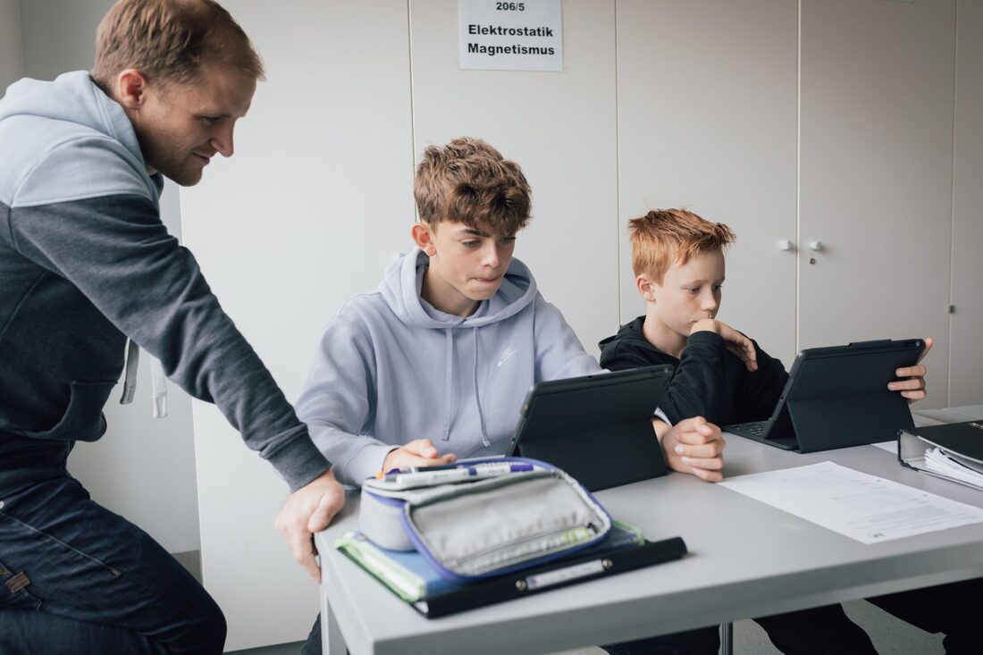 Ein Lehrer schaut zwei Schülern bei der Arbeit am iPad über die Schulter